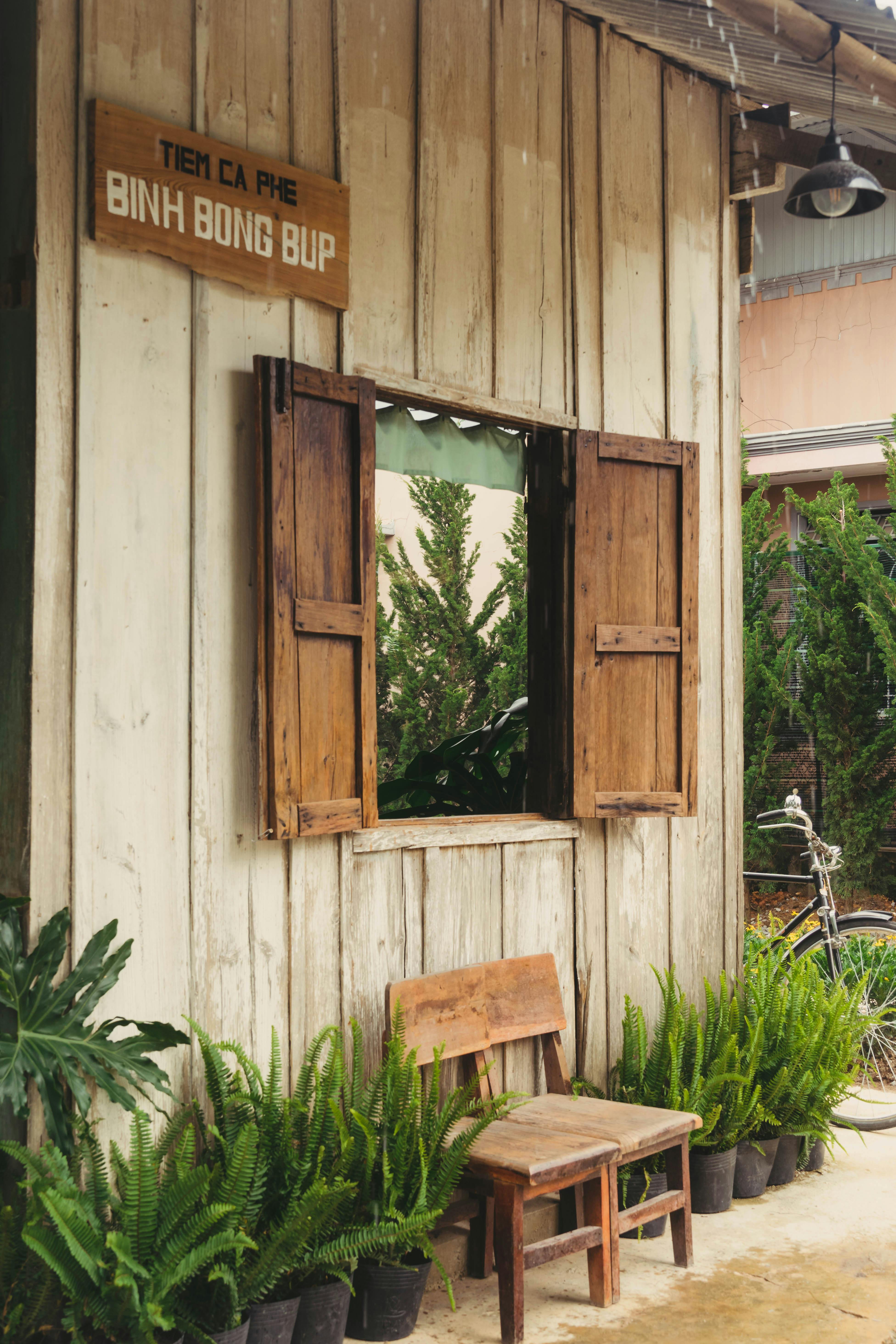 A charming wooden cafe with a "TIEM CA PHE BÌNH BÔNG BÚP" sign, open window, wooden chair, and lush green plants in pots, adding to the rustic and inviting atmosphere.